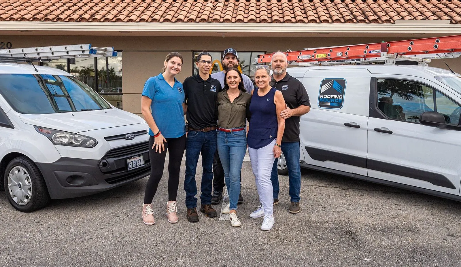 Six RoofingSource teammates standing together between two ladder-racked company vans outside a client restaurant.