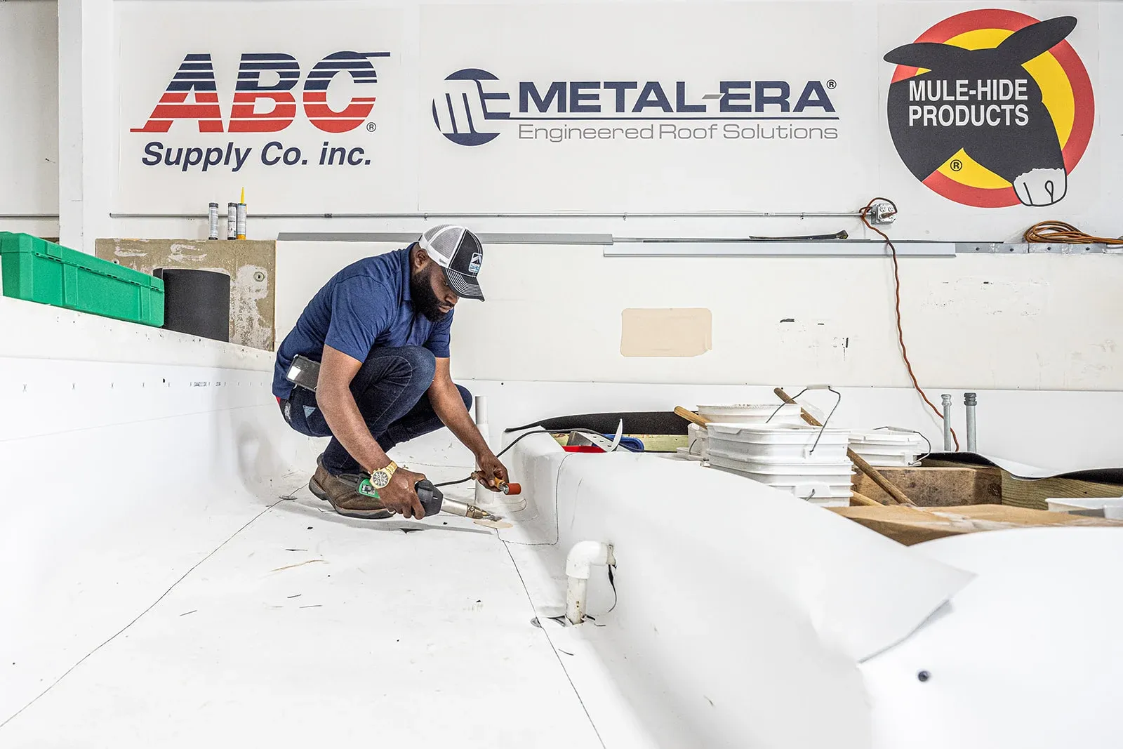 A teammate working on a roof deck in the training centre, manufacturer partner signs on the wall behind him.