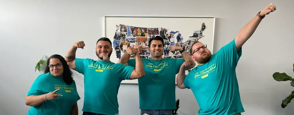 Four teammates in matching National Roofing Week shirts, arms raised, in front of the photo-collage map on the wall at headquarters.