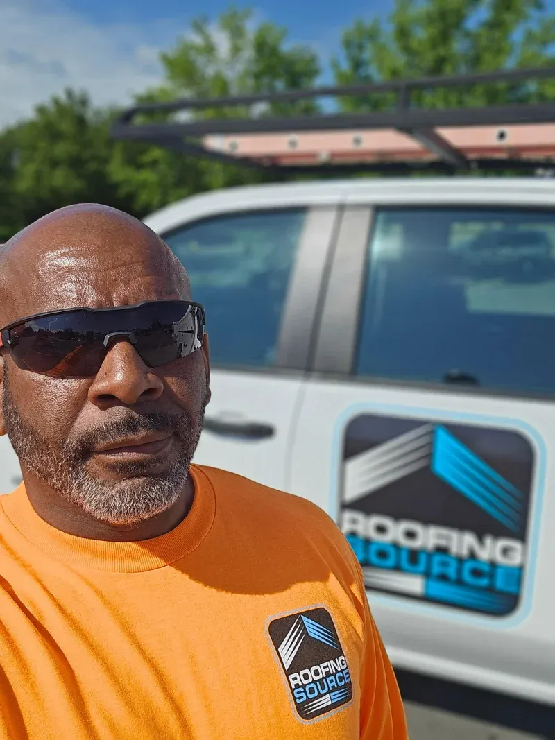 A Field Advisor in an orange RoofingSource shirt and sunglasses beside a company truck.