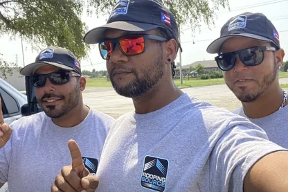 Three RoofingSource teammates in company caps and tees outside, one holding a finger up to the camera