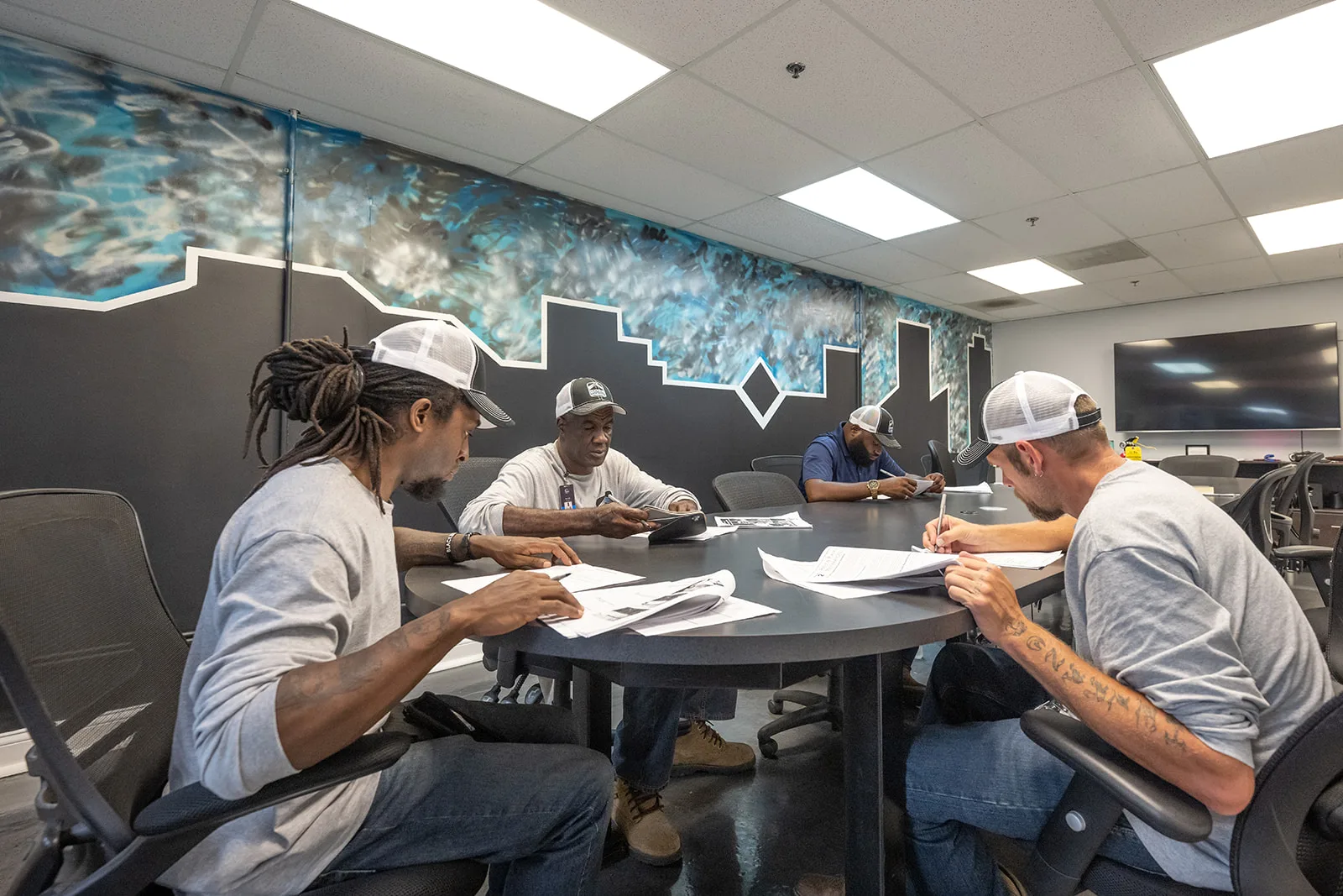 Four RoofingSource teammates around a table at headquarters, one pointing at a laptop while the others follow along.