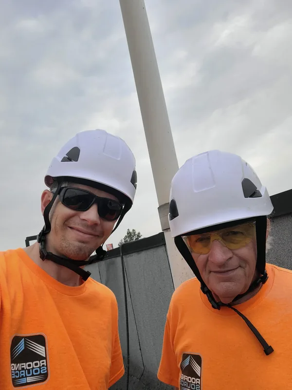 Two teammates in white hard hats and orange shirts, a selfie on a roof.