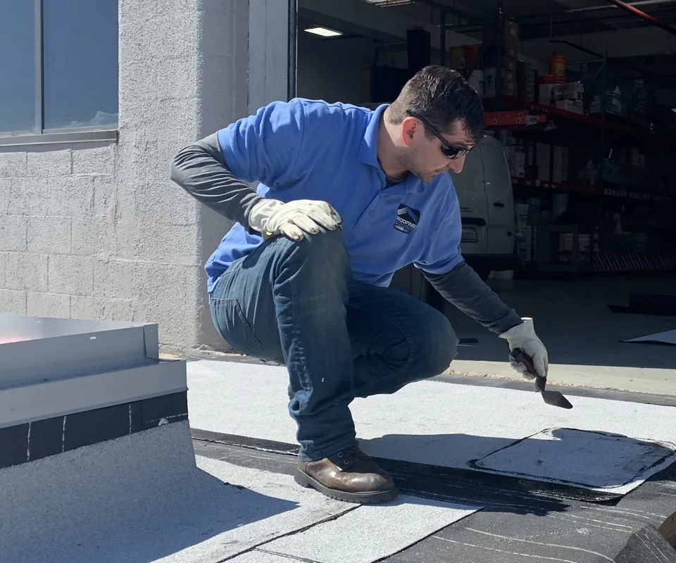 A teammate crouching over a roofing membrane with a trowel, a company van in the bay behind.