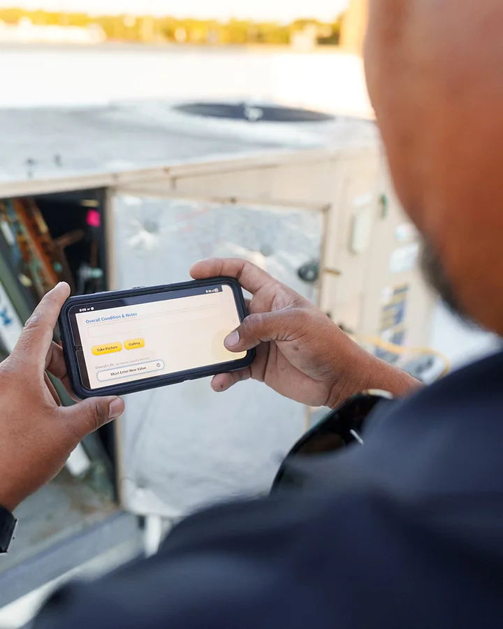 A teammate holding a phone with a roof condition report open on it, an opened rooftop unit behind