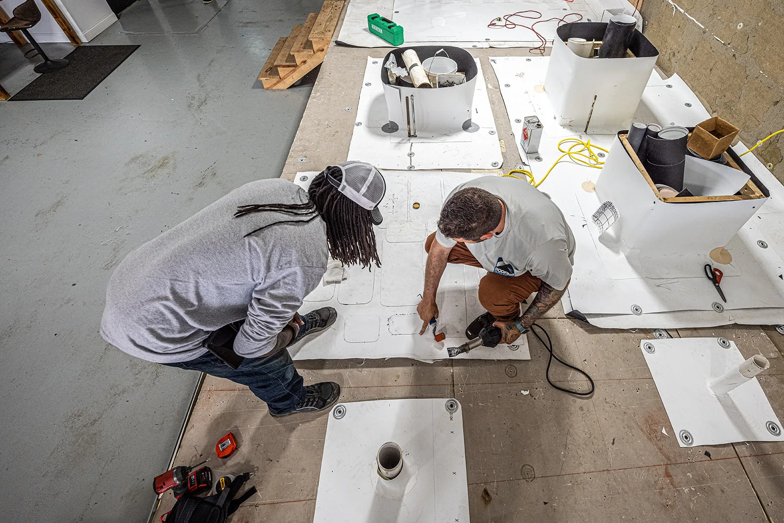 Seen from above, two teammates on a mock deck: one heat-welding a membrane patch around a pipe penetration while the other watches the work.