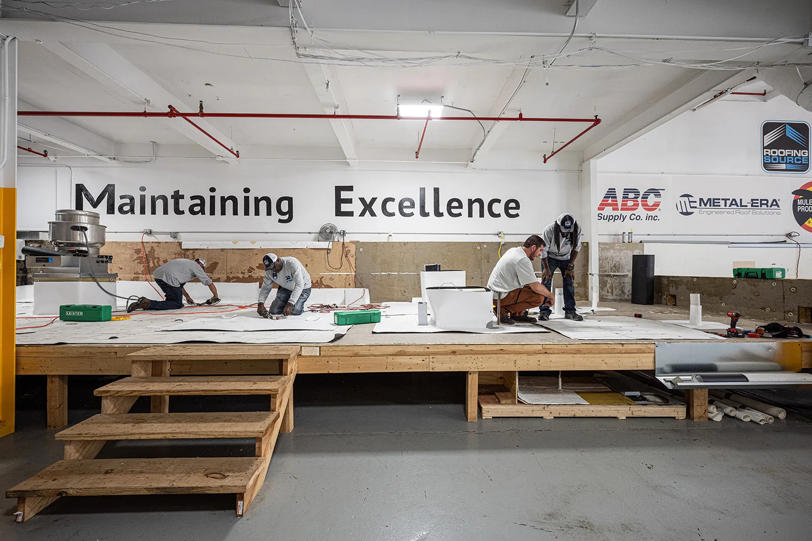 Four teammates at work on the indoor mock roof decks at headquarters, under a wall painted Maintaining Excellence.