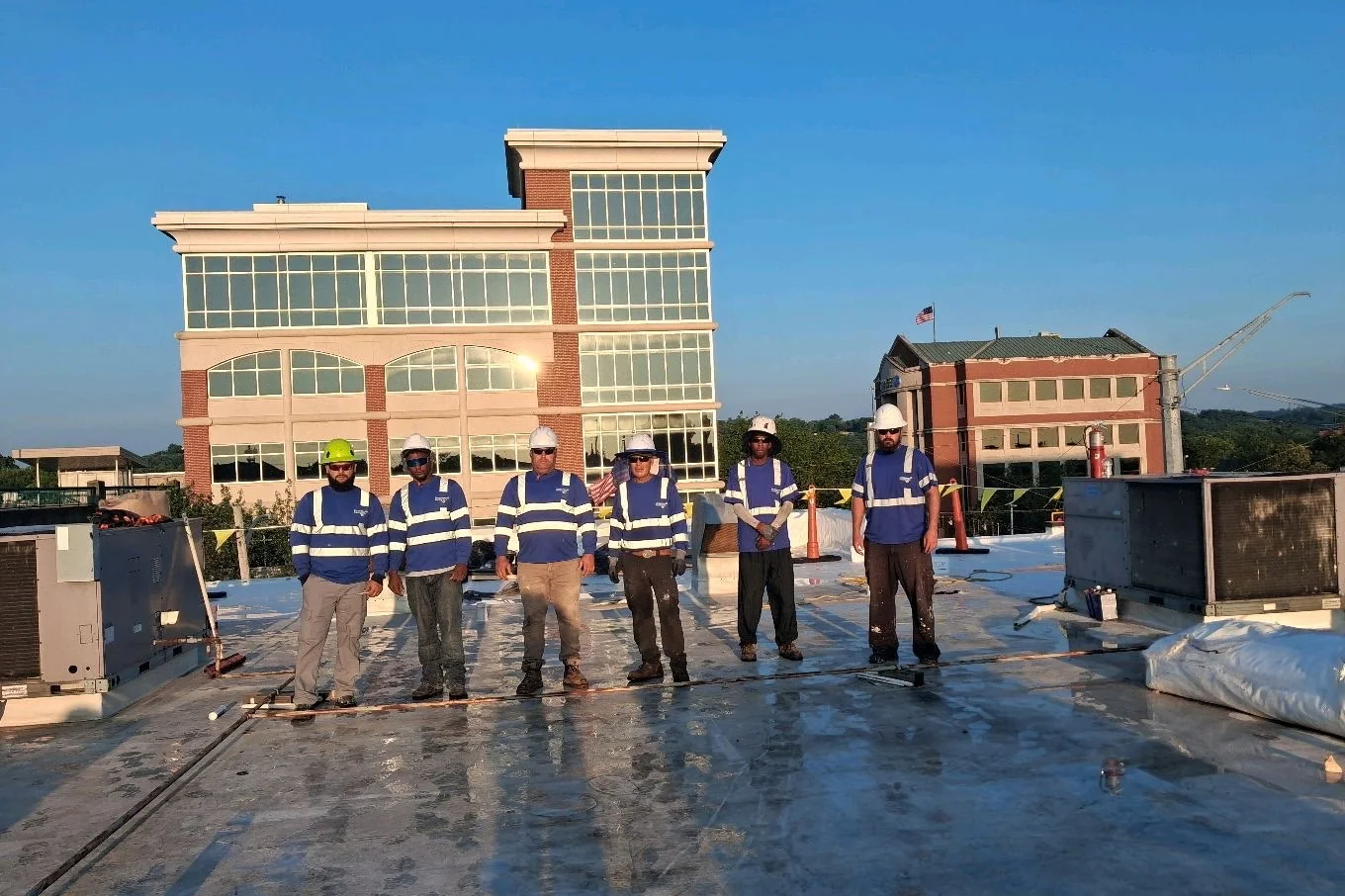 Six roofing crew members in blue hi-vis shirts and hard hats standing on a commercial rooftop at dusk, two brick office buildings behind them