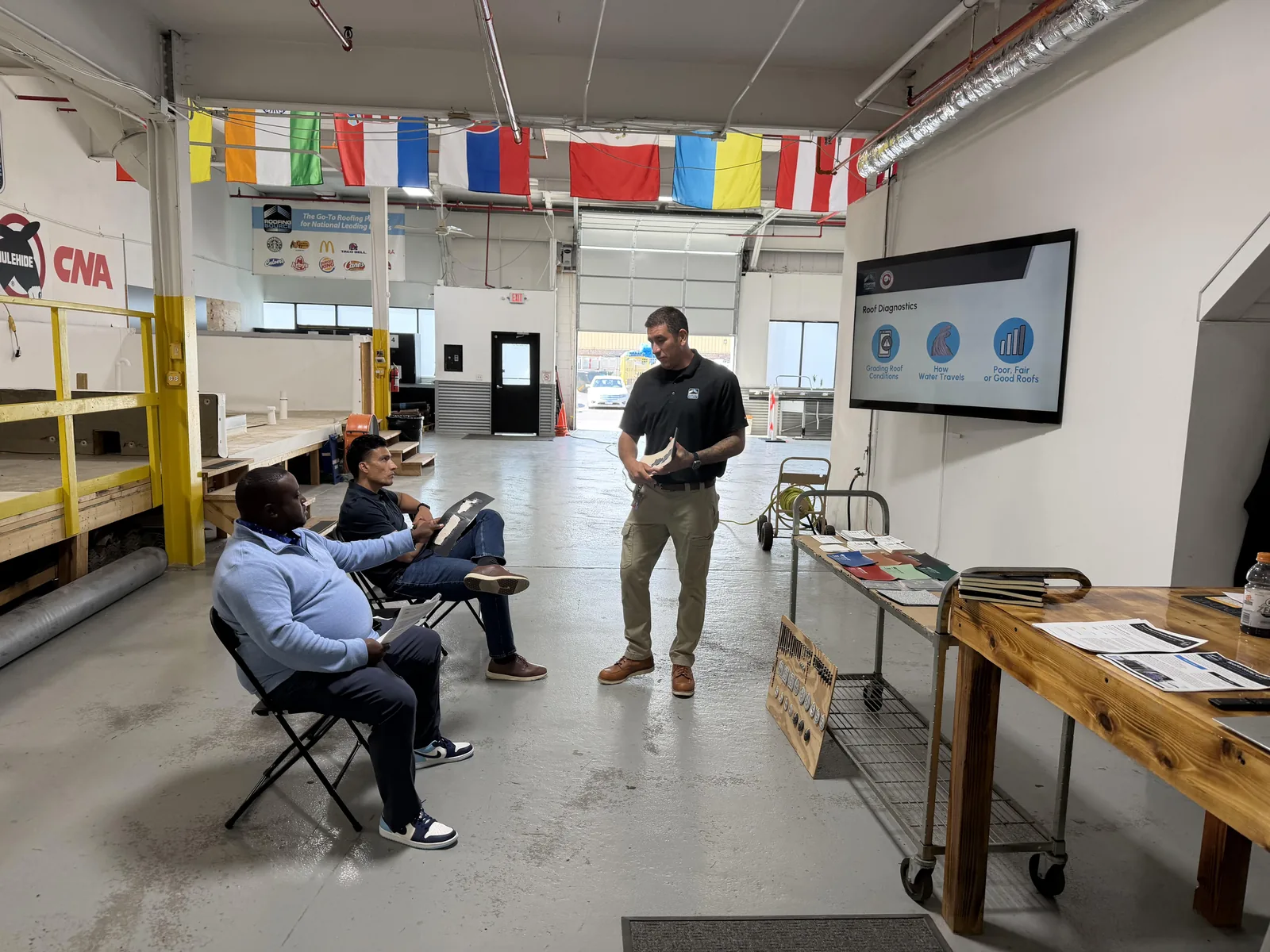 A RoofingSource teammate presenting roof diagnostics on a wall screen to two seated guests in the training bay, international flags overhead