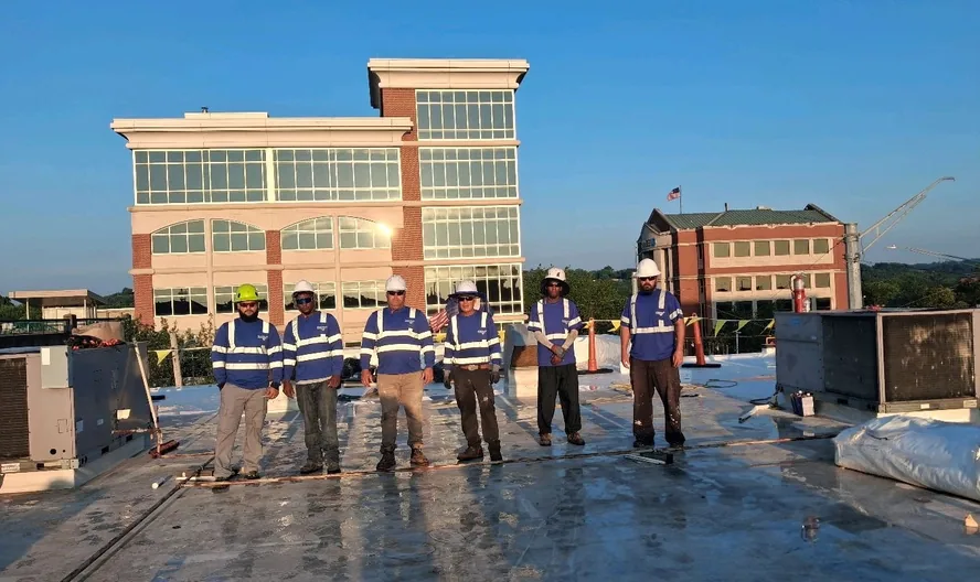 Six RoofingSource crew members standing in a line on a commercial roof at dusk, an office building behind them
