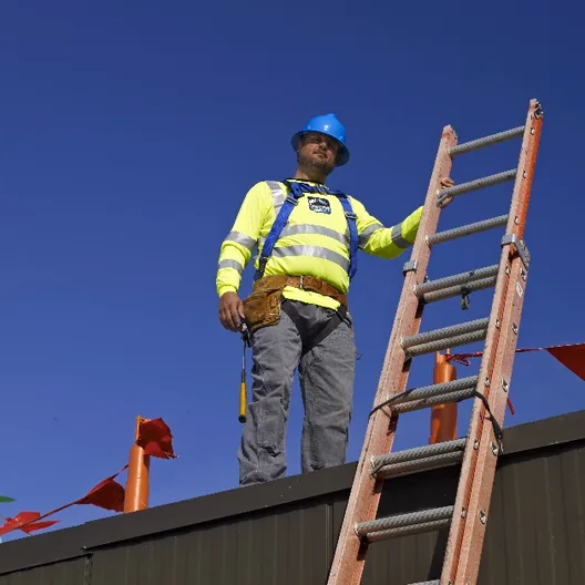 A roofer in a hard hat and harness at the top of a ladder against a blue sky