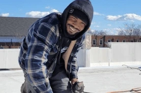 A teammate in a hooded winter jacket kneeling on a white roof membrane, smiling mid-repair