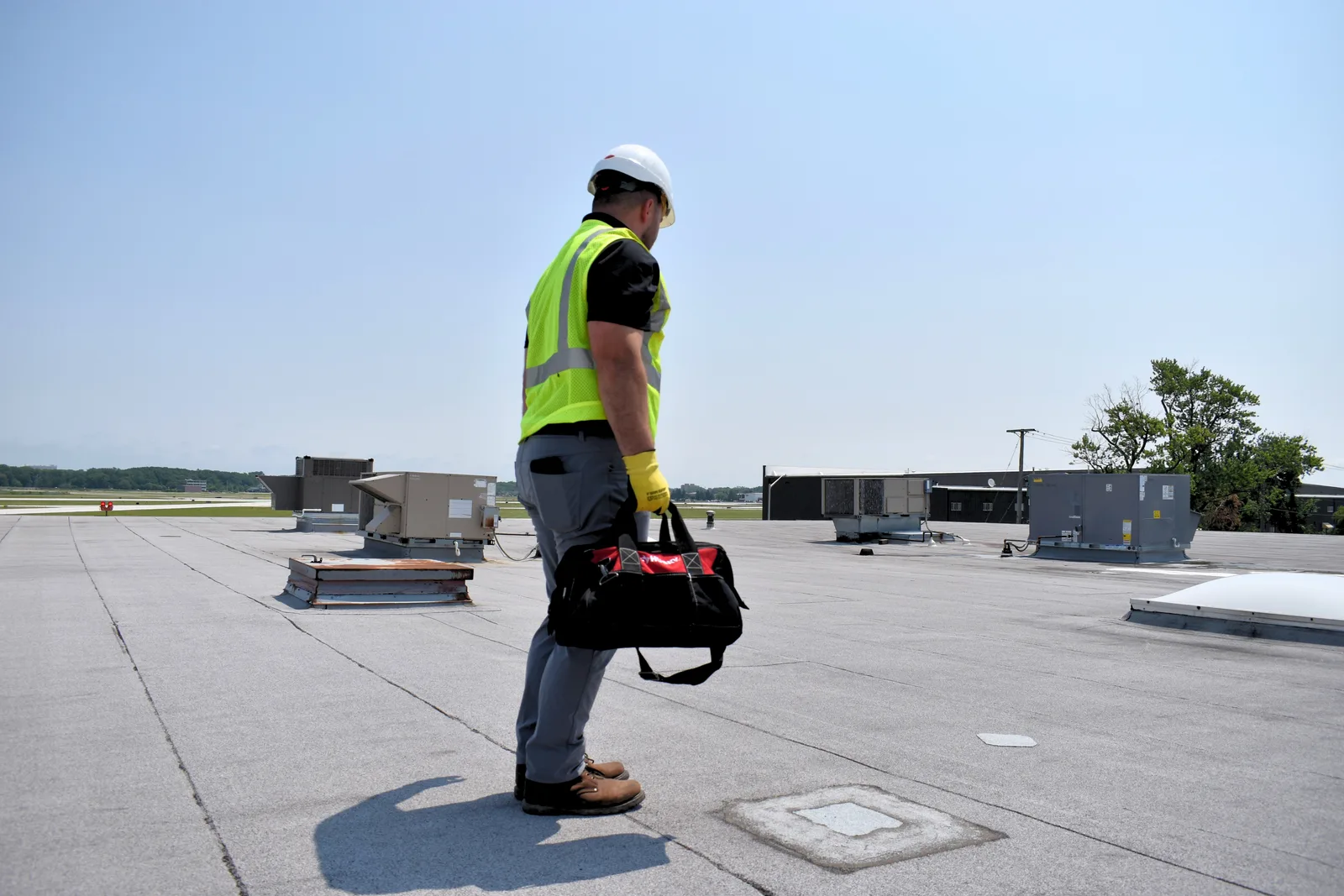 A RoofingSource teammate carrying a tool bag across a large commercial roof, rooftop units behind
